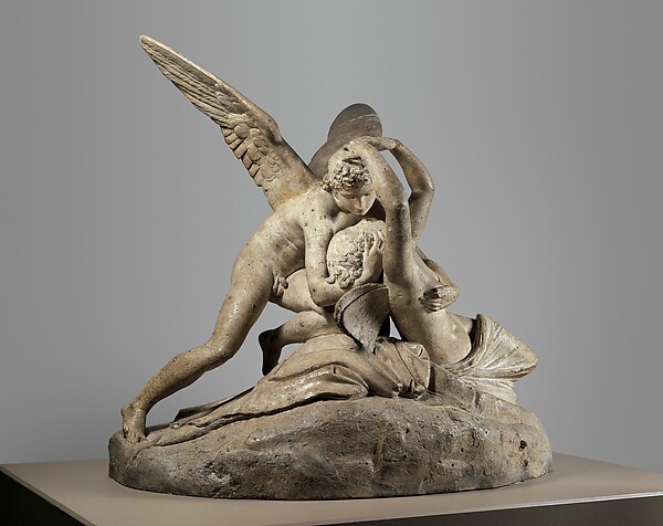 A winged Cupid leans over the reclining Psyche. Her raised arms curve around his head; their faces nearly meet. The weathered plaster rests on a rocky base.