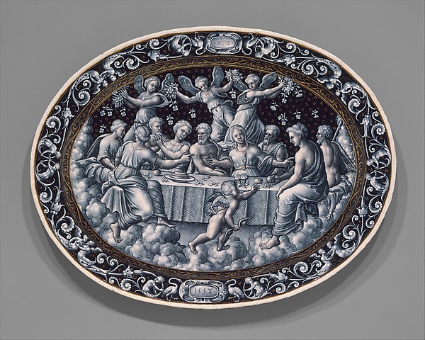 An oval enamel dish shows a crowded feast among clouds, painted in pale grey. Winged figures scatter flowers above the table; scrolling ornament and gold surround the scene.